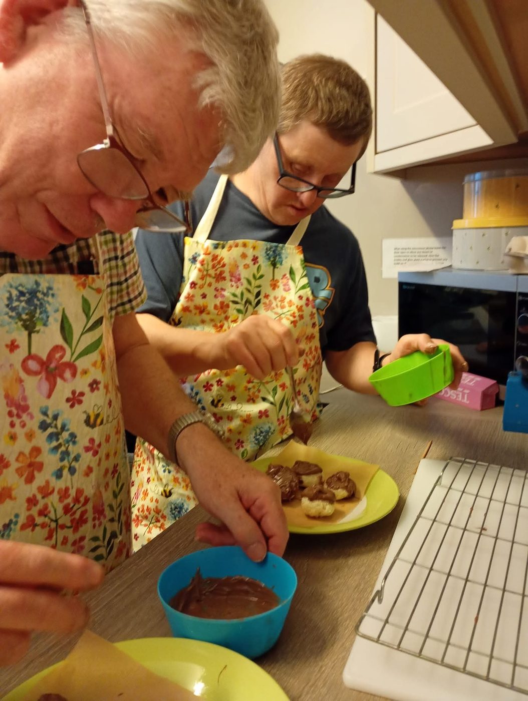 Two people in floral aprons dipping treats in chocolate at the kitchen worktop.