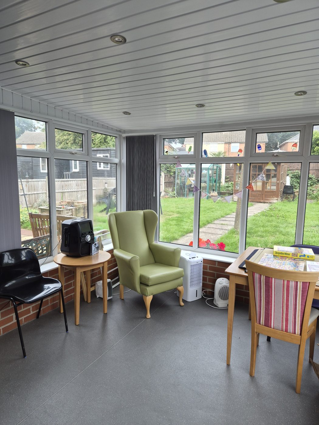 A bright conservatory with a green armchair, a jigsaw table and bunting, looking out to the garden.