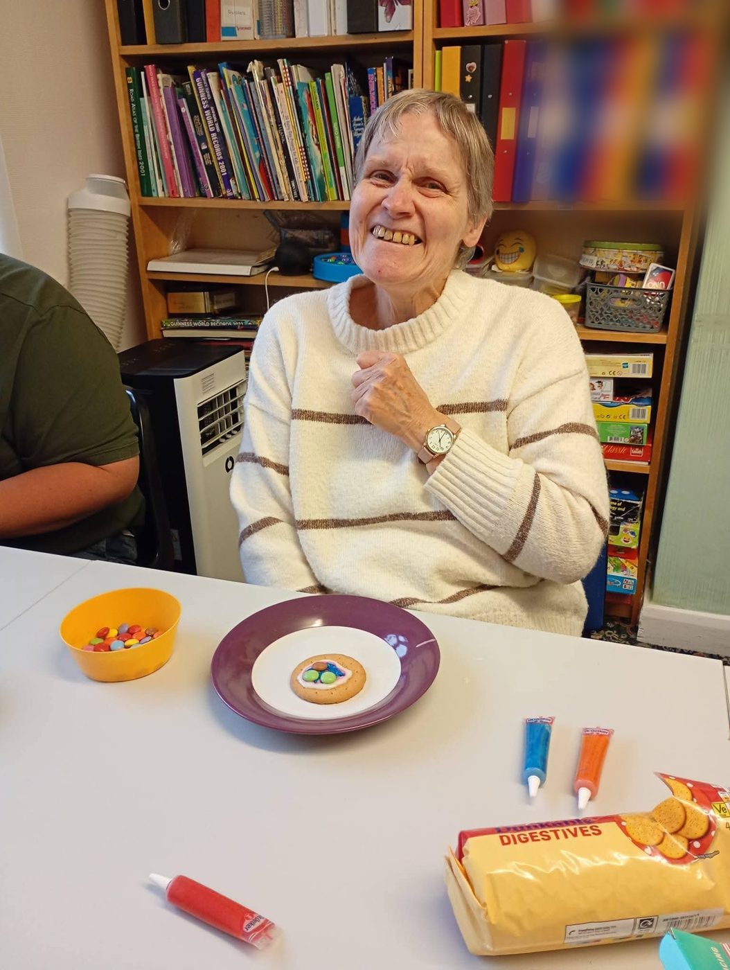 A woman laughing at a table beside a biscuit she has decorated with icing and sweets.
