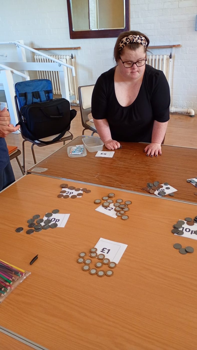 A woman sitting at a table with piles of coins sorted onto labelled cards, practising counting money.