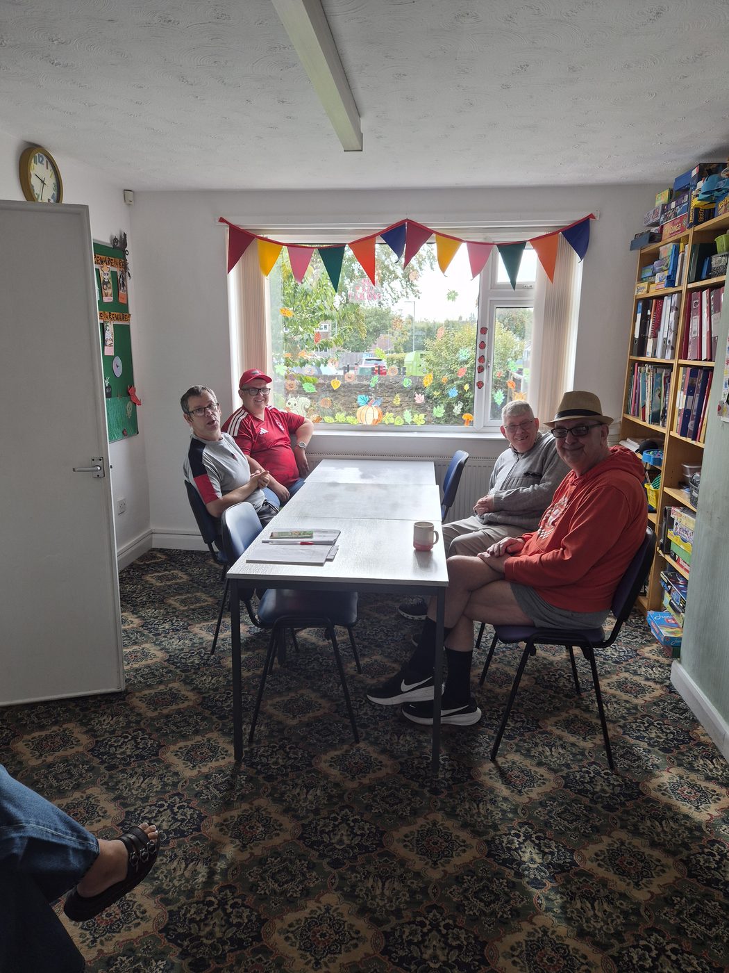 Four friends smiling around a table in a room with rainbow bunting and shelves of games.