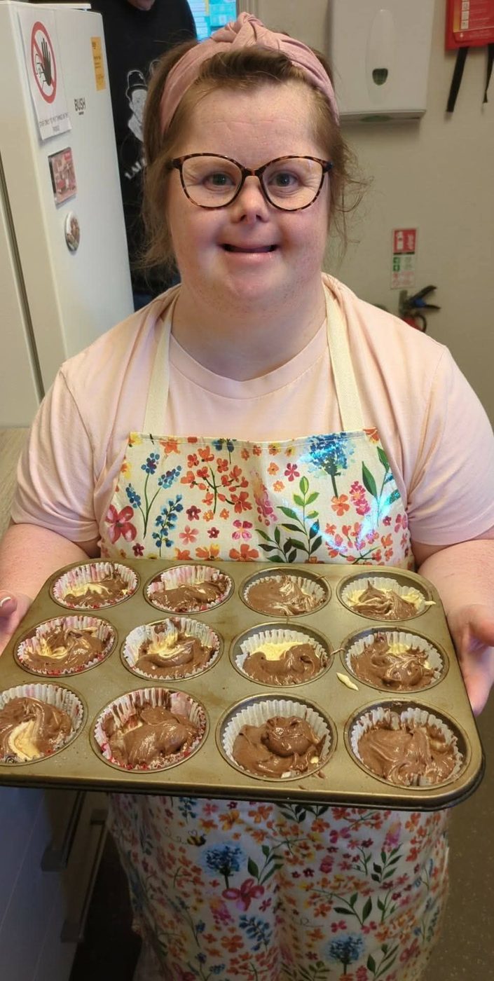 A smiling woman in an apron holding a tray of chocolate swirl muffins ready to bake.