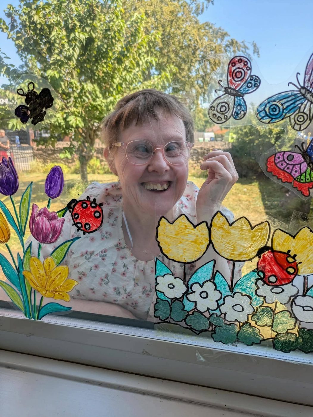 A smiling woman looking through a window decorated with hand-painted flowers, butterflies and ladybirds.